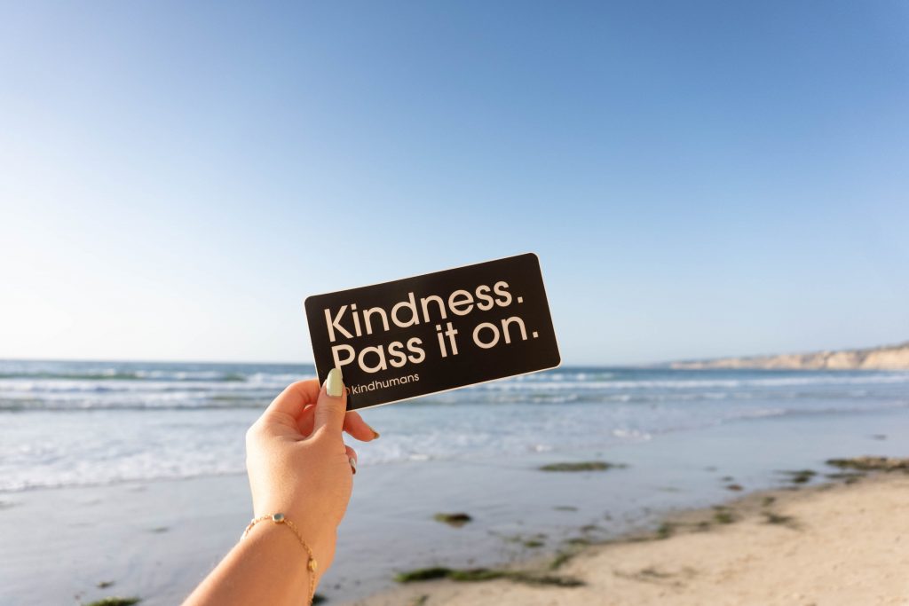 CommunityAd Trades - Charities - acts of kindness - person holding a black notepad that says 'Kindness. Pass it on.' in front of beach